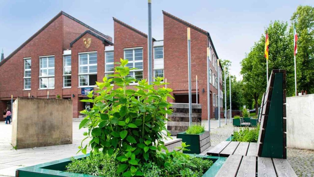 Grüne Pflanzen in Hochbeeten vor einem modernen, roten Backsteingebäude mit Flaggen.