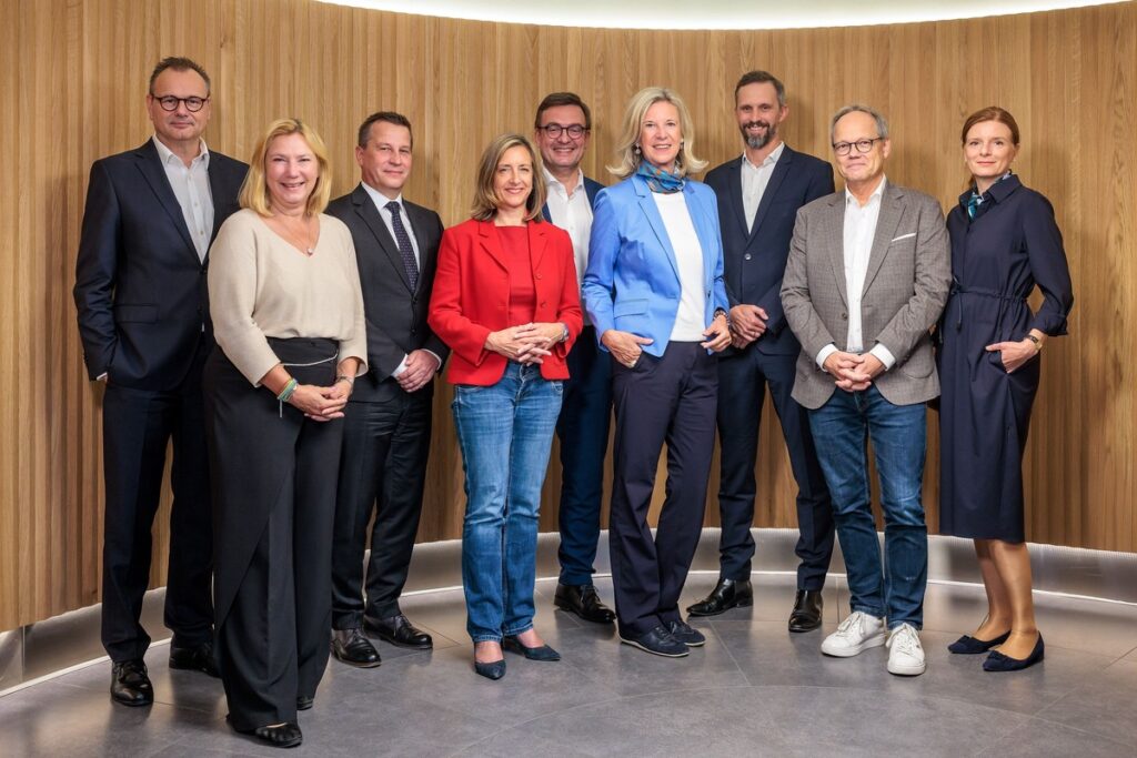 Eine Gruppe von neun Personen in formeller Kleidung, die in einem modernen Innenraum aufgestellt sind.