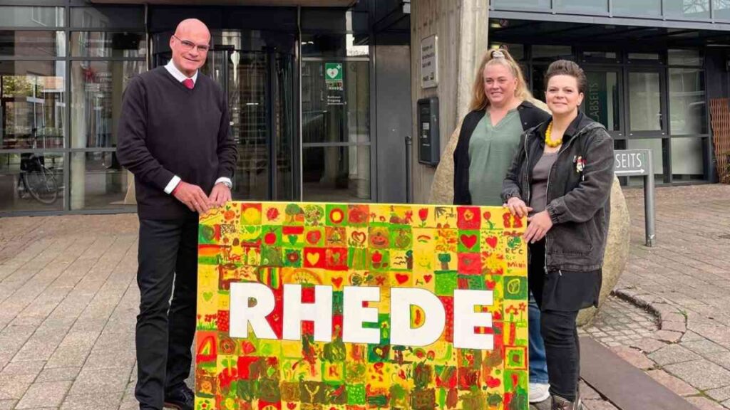 Drei Personen stehen vor einem Gebäude und halten ein buntes Plakat mit der Aufschrift 'RHEDE'.