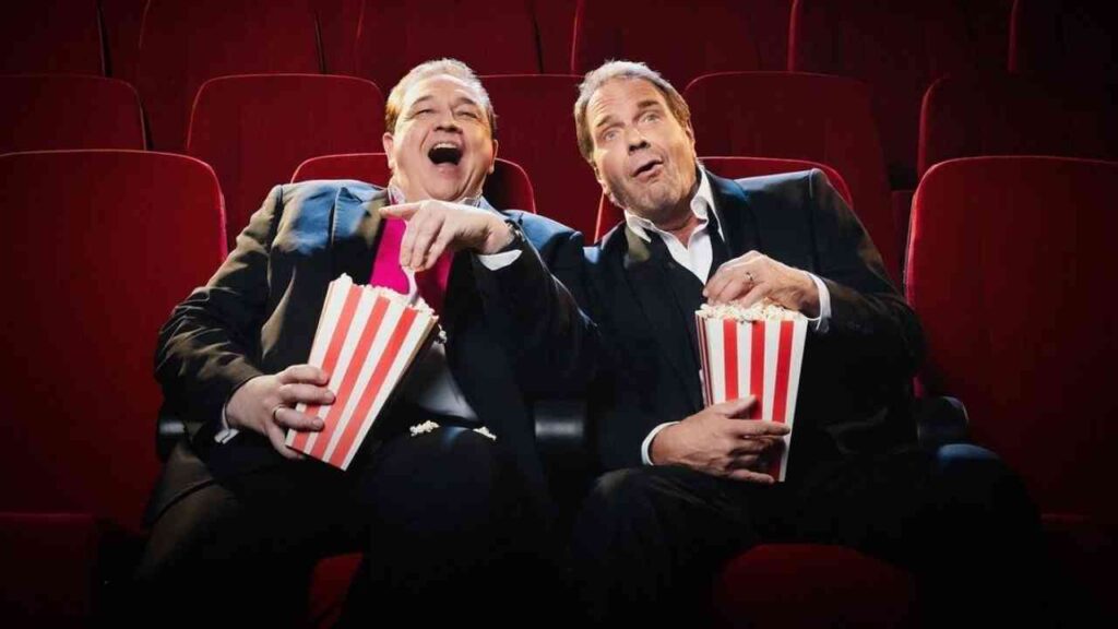 Zwei Männer in Anzügen sitzen im Kino und essen Popcorn aus roten und weißen Tüten.