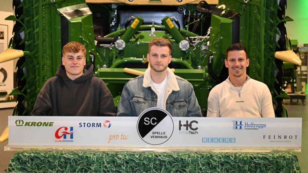 Drei Personen sitzen an einem Tisch mit einem Vertrag, während ein Traktor im Hintergrund sichtbar ist.
