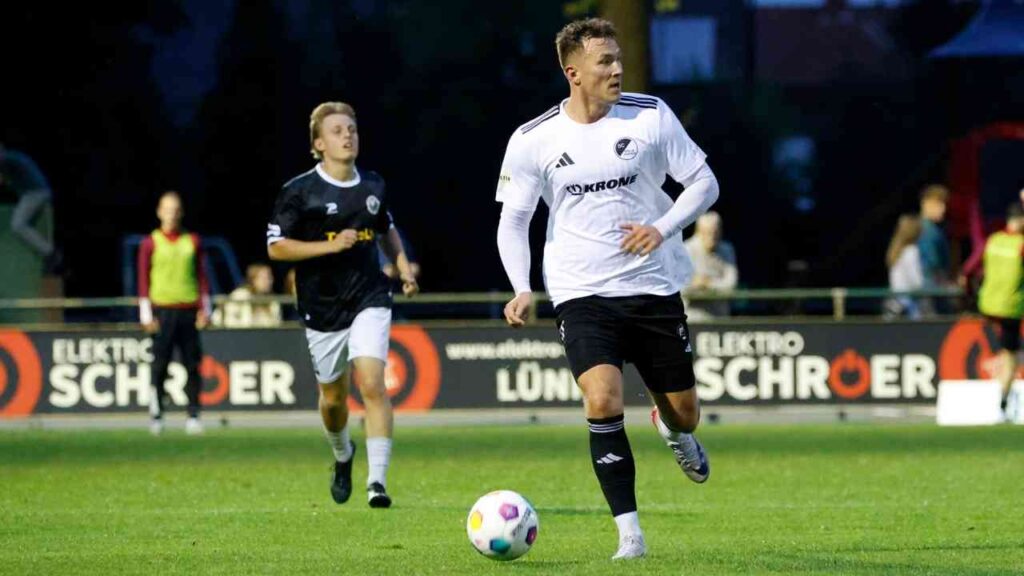 Fußballspieler in weißem Trikot mit schwarzer Hose, der den Ball auf dem Spielfeld führt.