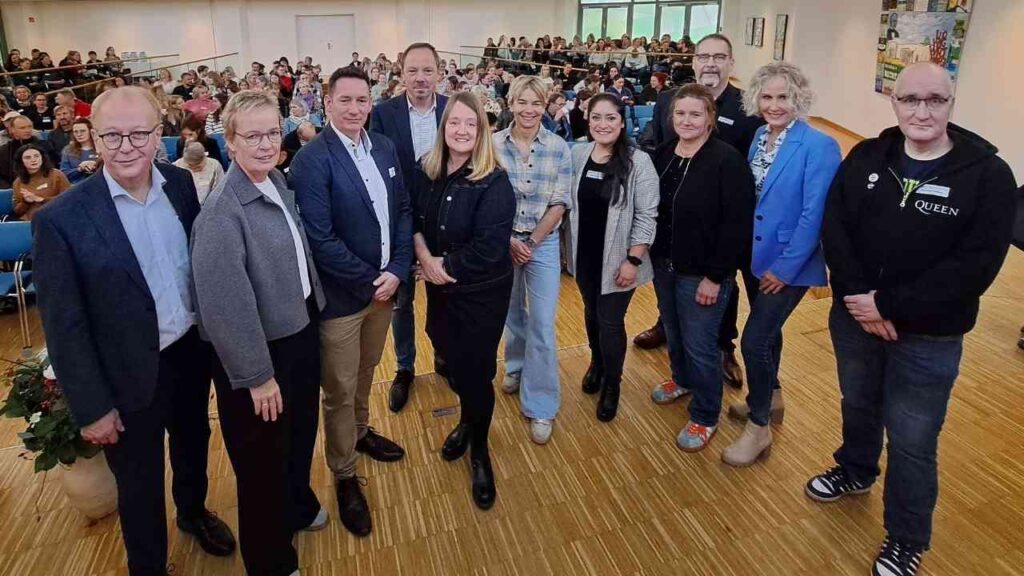 Gruppe von Personen, die in einem Raum für eine Veranstaltung stehen, mit Fokus auf die zentrale Person in schwarzer Kleidung.