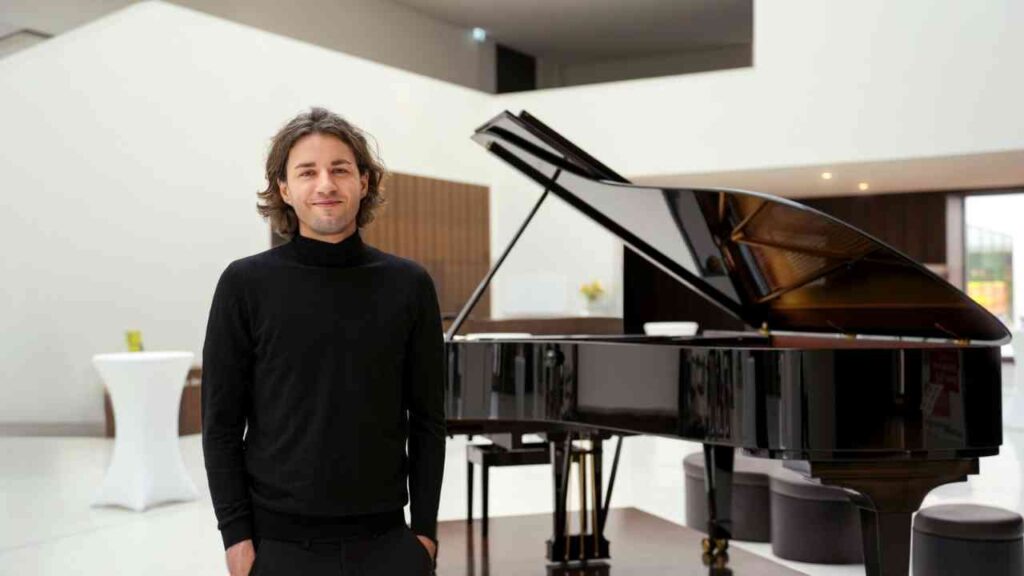 Mann in schwarzem Pullover steht neben einem schwarzen Flügelpiano in einem modernen Raum.