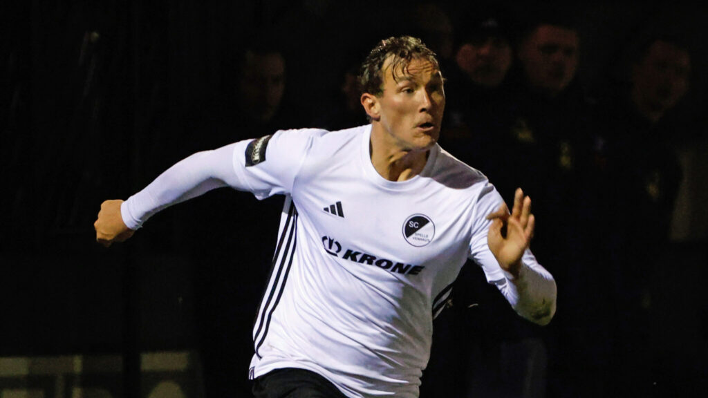 Fußballspieler in einem weißen Trikot mit schwarzem Logo, der während eines Spiels sprintet.