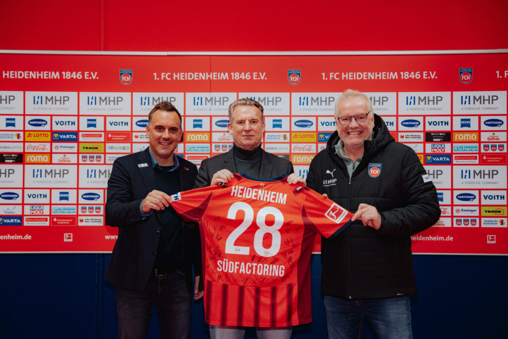 Drei Personen halten ein signiertes Trikot des 1. FC Heidenheim mit der Nummer 28 in der Hand.