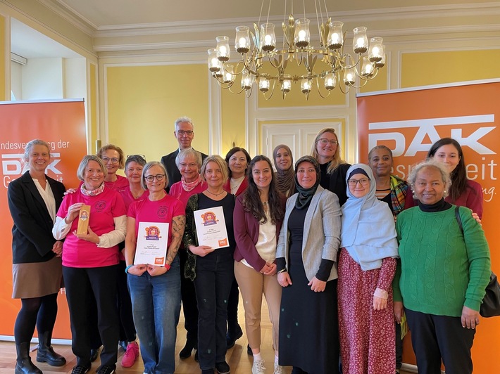 Gruppe von Personen in pinken T-Shirts, die Auszeichnungen und Urkunden in einem Raum mit PAK-Logos halten.