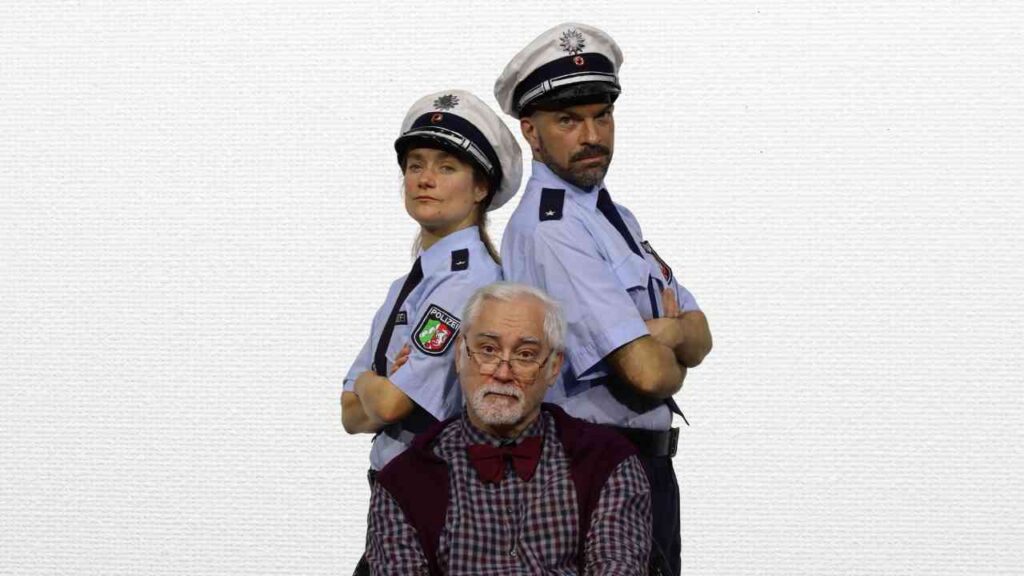 Zwei Polizisten in Uniform stehen hinter einem älteren Mann mit Brille und einem roten Pullover.