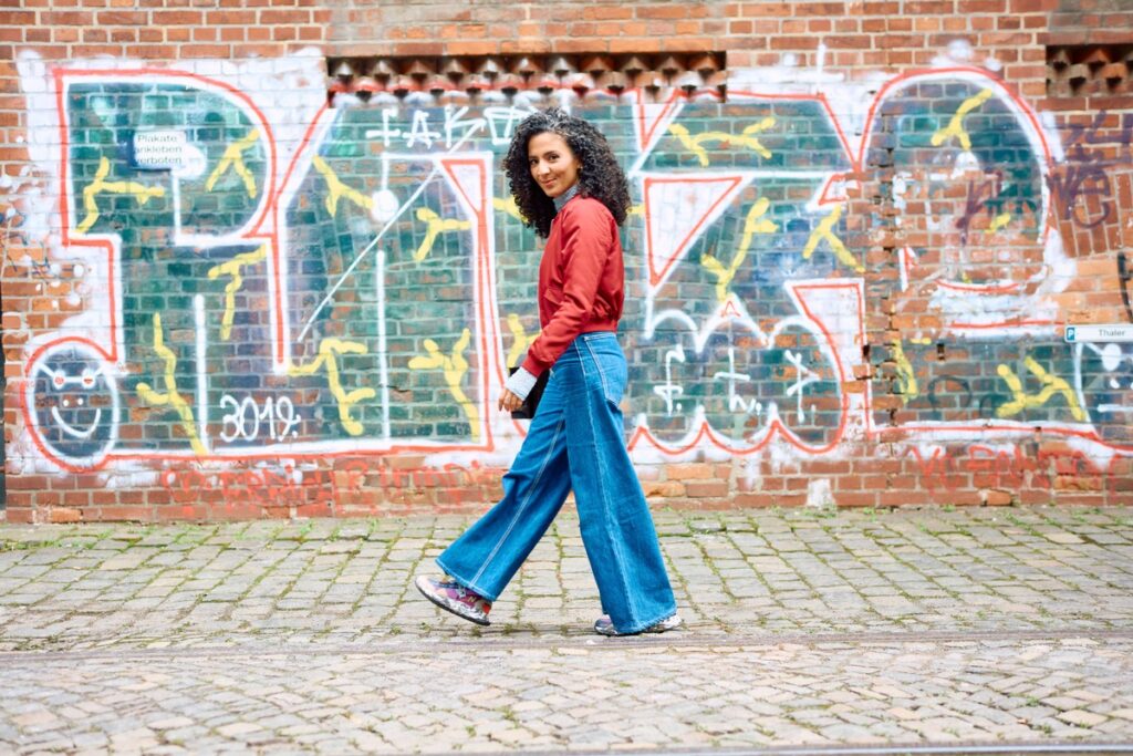 Frau mit lockigem Haar in rotem Oberteil und blauen Jeans, die an einer mit Graffiti bemalten Wand entlanggeht.