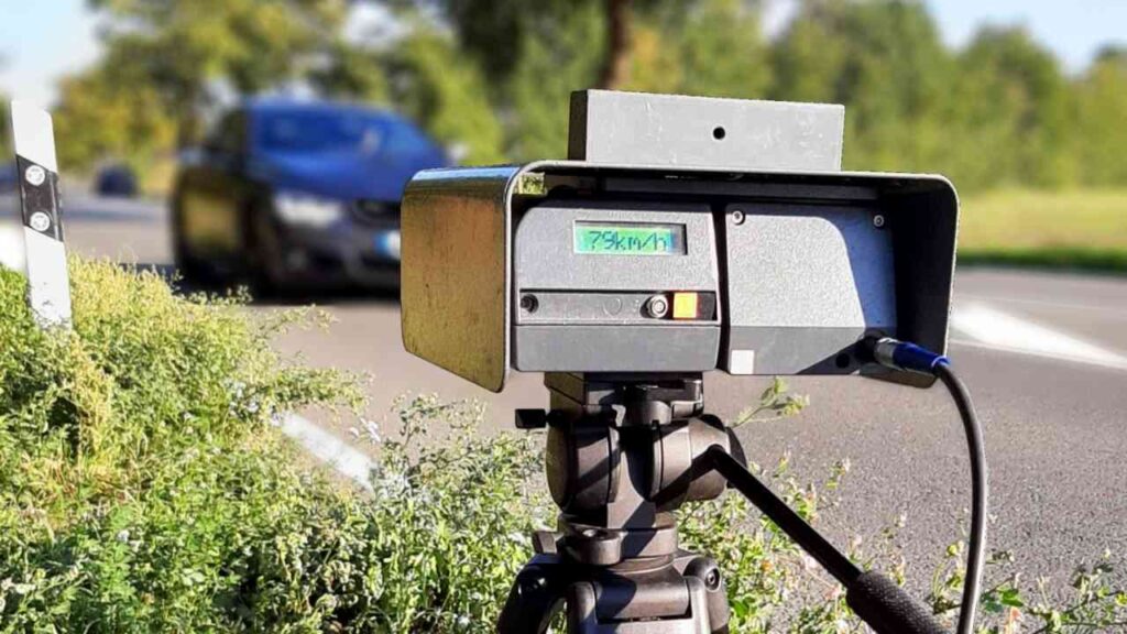 Geschwindigkeitsmessgerät auf einem Stativ an einer Straße, mit einem vorbeifahrenden Auto im Hintergrund.
