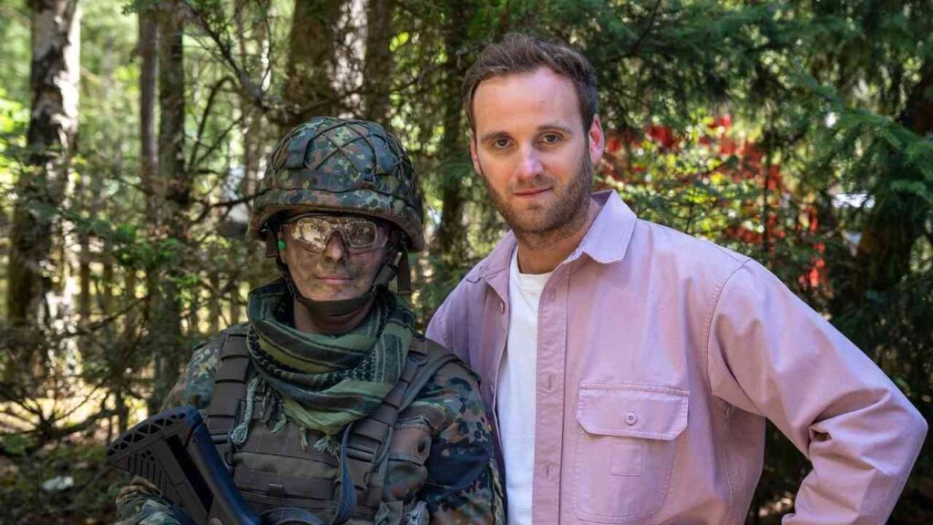 Ein Soldat in Tarnkleidung mit Gesichtsbemalung steht neben einem Mann in einem rosa Hemd im Wald.