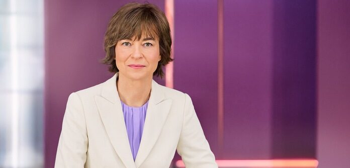 Frau mit kurzen, braunen Haaren in einem hellen Blazer und lila Bluse, posiert vor einem lila Hintergrund.