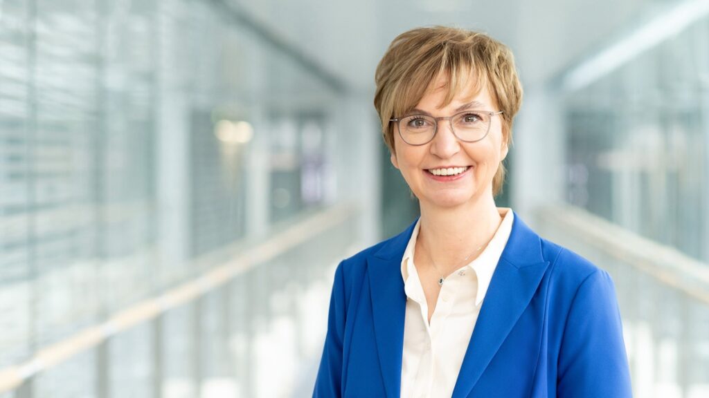 Ursula Weyrich in einem blauen Blazer mit Brille, lächelnd vor einem modernen Hintergrund.