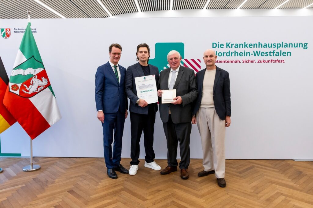 Vier Personen stehen zusammen und halten eine Auszeichnung in der Hand, im Hintergrund ein Banner zur Krankenhausplanung Nordrhein-Westfalen.