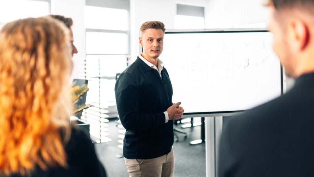 Mann in schwarzem Pullover präsentiert Informationen vor einer Gruppe in einem modernen Büro.