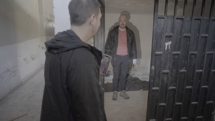 Zwei Männer in einem Raum mit Gittertür, einer hält ein Smartphone und schaut den anderen an.