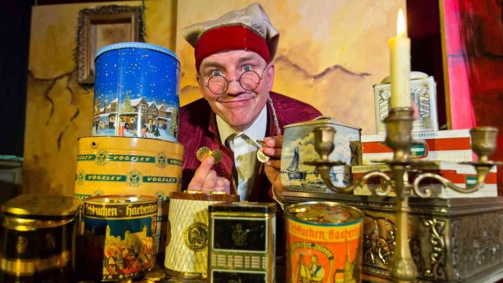 Ein Mann in historischer Kleidung zeigt eine Münze vor einer Sammlung von alten Dosen und einem Kerzenständer.