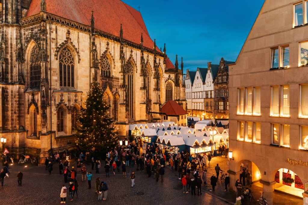 Weihnachtsmarkt in Münster mit festlich beleuchteten Ständen und einer großen Tanne vor der Kirche bei Dämmerung.