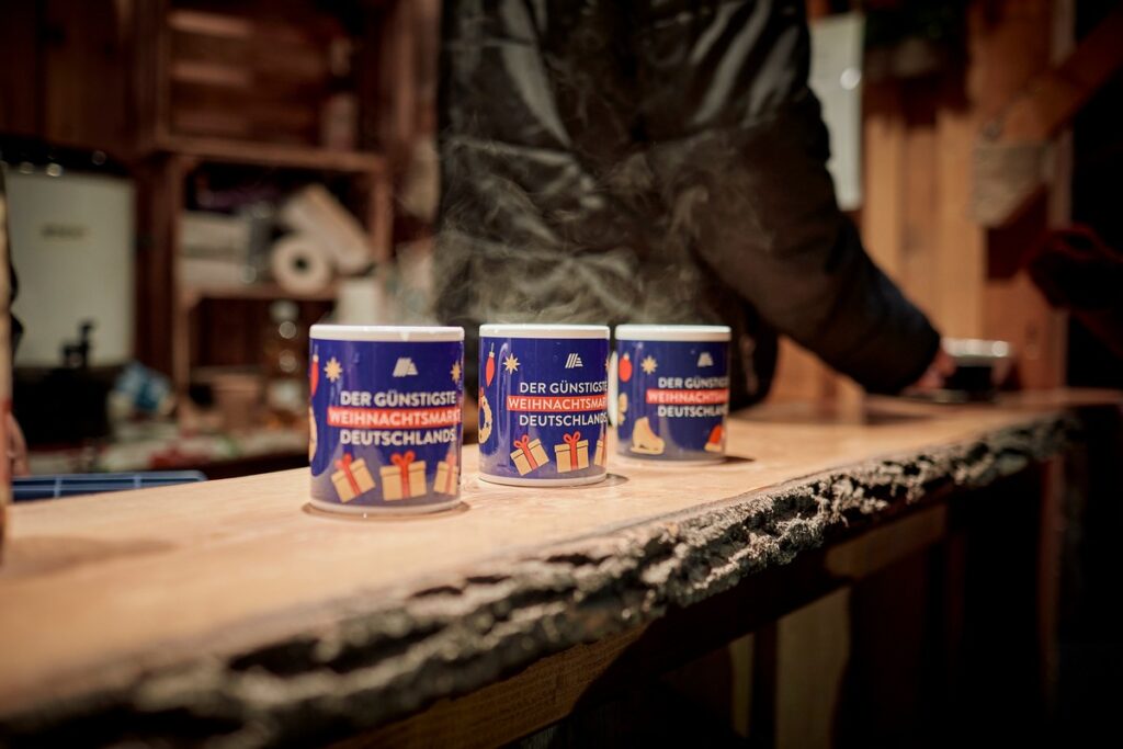 Drei Tassen mit dem Aufdruck 'Der günstigste Weihnachtsmarkt Deutschlands' auf einem Tisch.