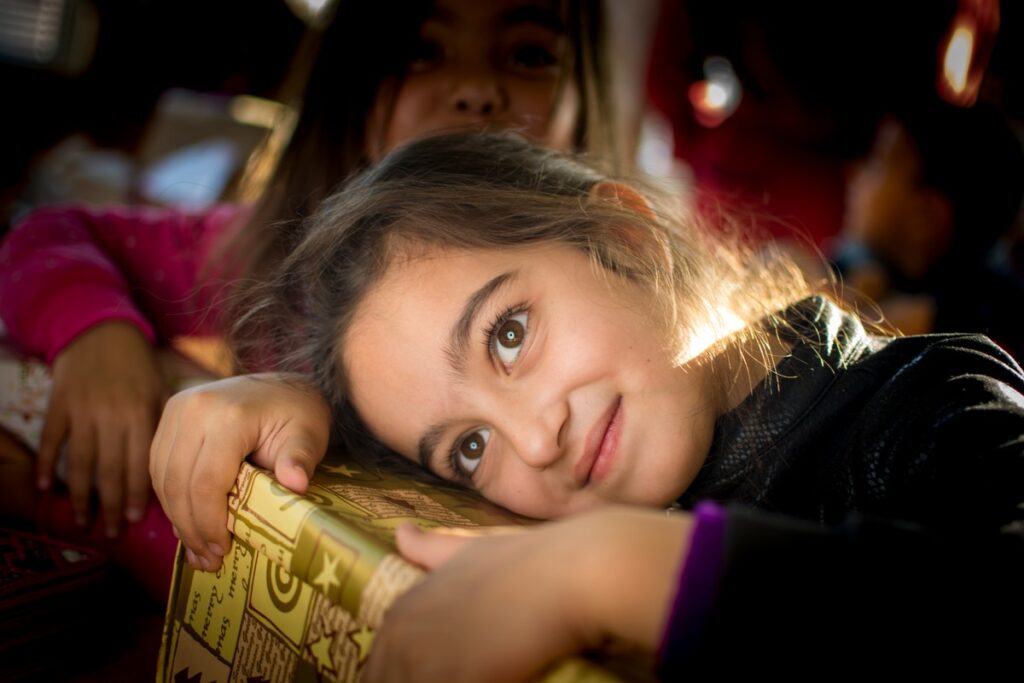 Ein Mädchen mit braunen Augen umarmt ein Geschenk mit goldener Verpackung und lächelt in die Kamera.