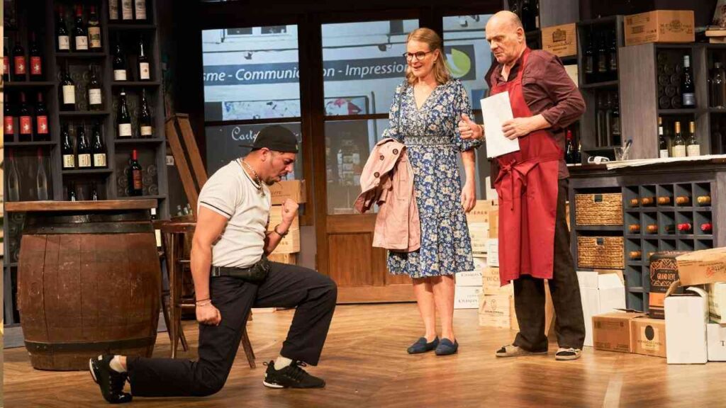 Szenenbild einer Theateraufführung mit drei Darstellern in einem Weingeschäft, einer kniet auf dem Boden.