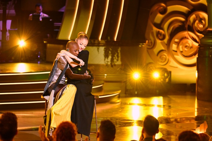 Zwei Künstlerinnen umarmen sich auf einer Bühne während einer musikalischen Veranstaltung mit Beleuchtung im Hintergrund.