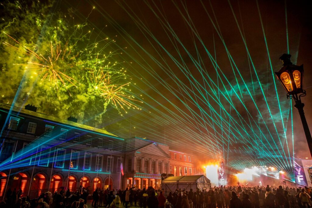 Lichtshow mit Laserstrahlen und Feuerwerk über einer Menschenmenge in einer urbanen Umgebung.