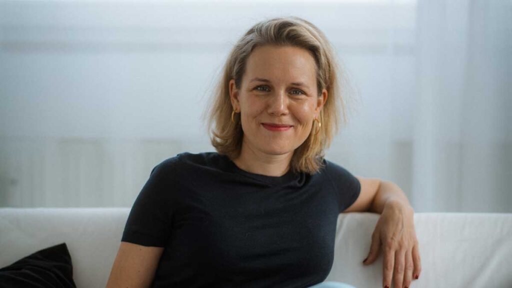 Frau mit blonden Haaren und lächelndem Gesicht, sitzt auf einer Couch in einem hellen Raum.