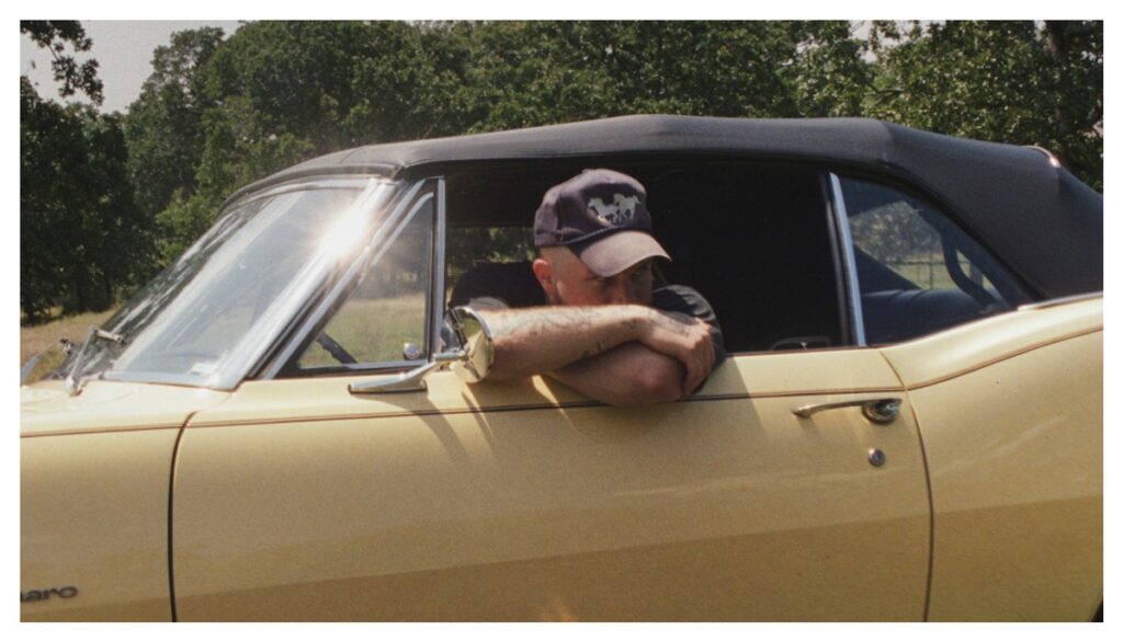 Mann mit einer Baseballmütze lehnt mit verschränkten Armen aus einem gelben Cabrio-Fenster.