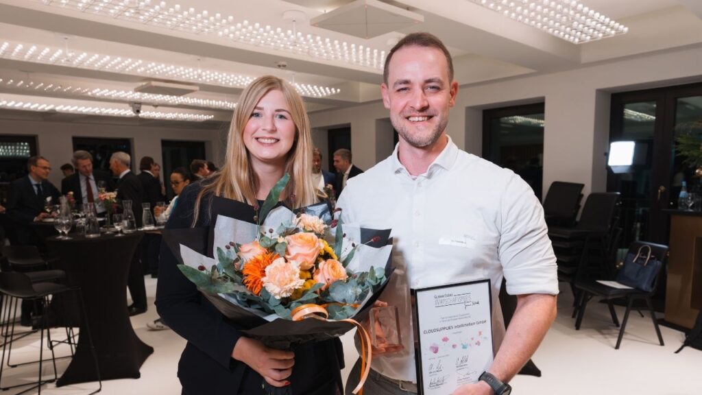 Zwei Personen halten einen Preis und einen Blumenstrauß bei einer Preisverleihung.