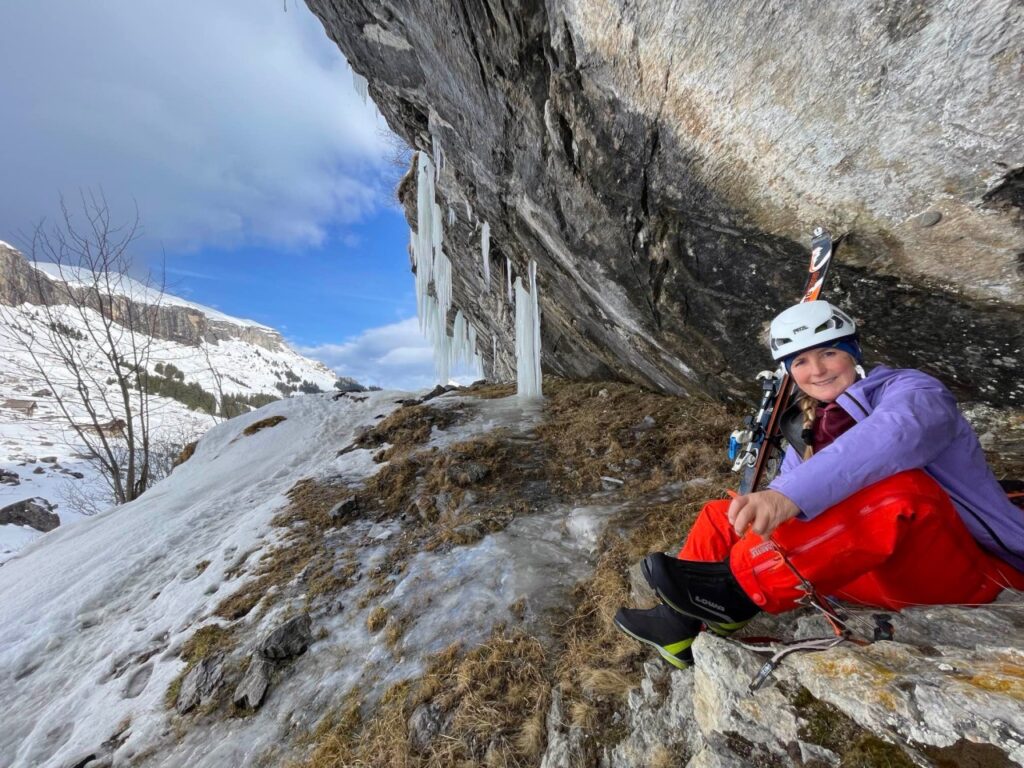 Eine Person in Kletterausrüstung sitzt auf einem Felsen unter einer steilen Felswand mit Eiszapfen.