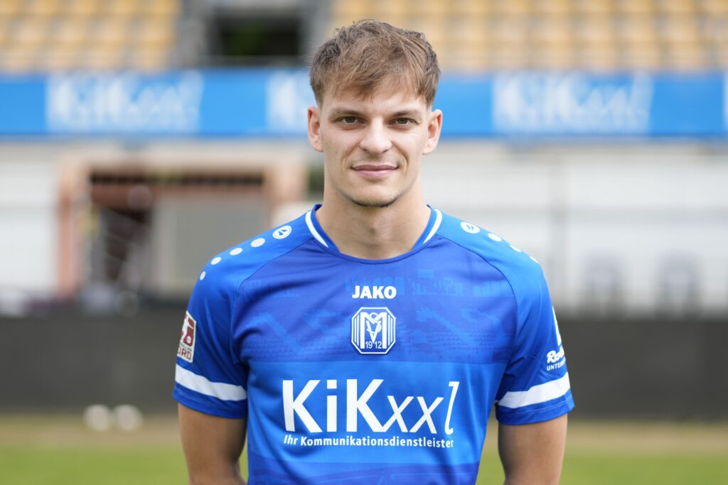 Ein junger Fußballspieler in einem blauen Trikot mit dem Logo eines Sponsors auf der Brust und einem neutralen Hintergrund.