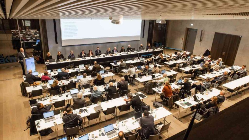 Konferenzraum mit Teilnehmern, die an Tischen sitzen und an einer Präsentation teilnehmen.