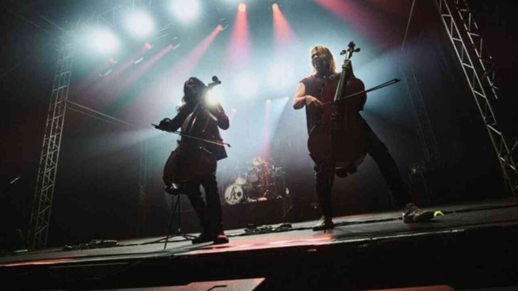 Zwei Cellisten auf der Bühne während eines Konzerts mit Scheinwerferlicht und einer Band im Hintergrund.