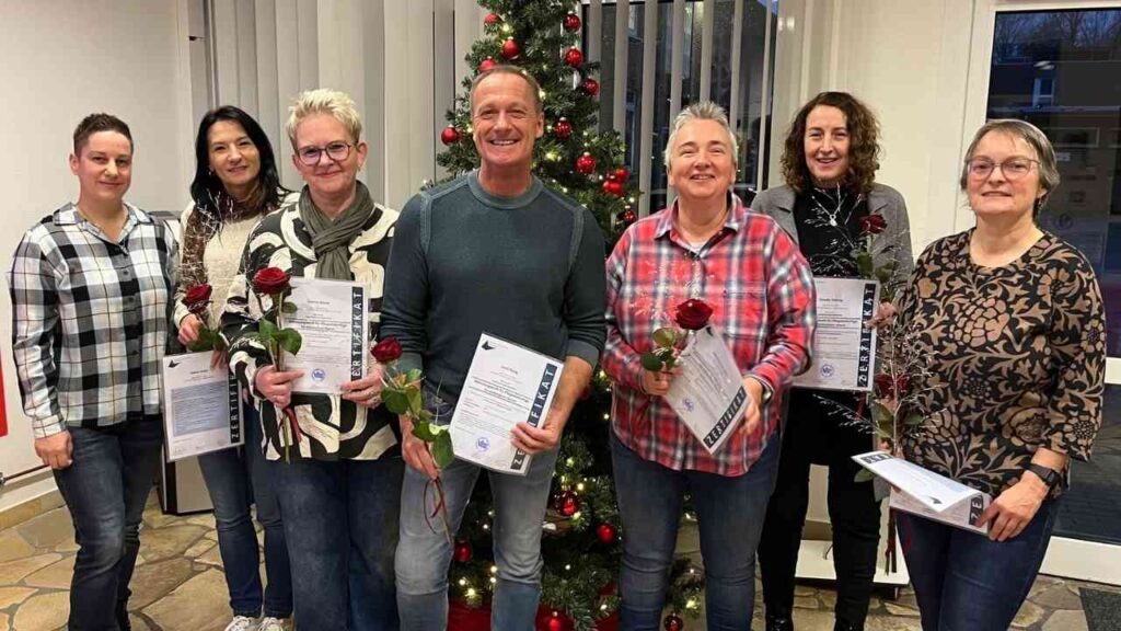 Gruppe von Personen, die stolz ihre Zertifikate in der Hand halten, mit roten Rosen, vor einem Weihnachtsbaum.
