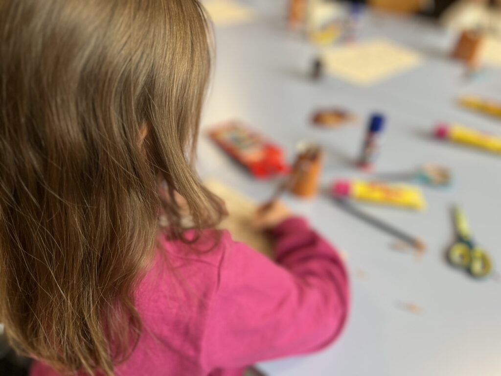 Ein Kind mit langen, braunen Haaren, das an einem Tisch mit Bastelmaterialien arbeitet.