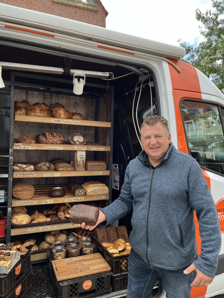 Mann hält ein Laib Brot vor einem Verkaufsstand mit verschiedenen Brotsorten in einem Verkaufswagen.