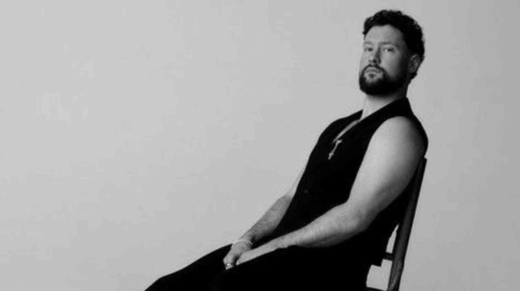 Calum Scott sitzt auf einem Stuhl, trägt ein ärmelloses Oberteil und hat einen Bart.