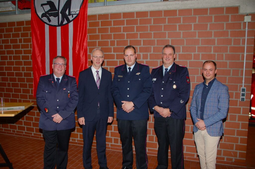 Fünf Männer in Uniformen und Anzügen stehen nebeneinander in einem Innenraum mit einer roten Fahne im Hintergrund.