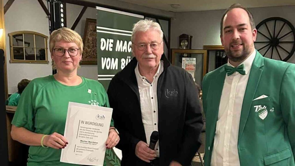 Drei Personen stehen zusammen und halten eine Urkunde in der Hand, während sie in einem Innenraum posieren.