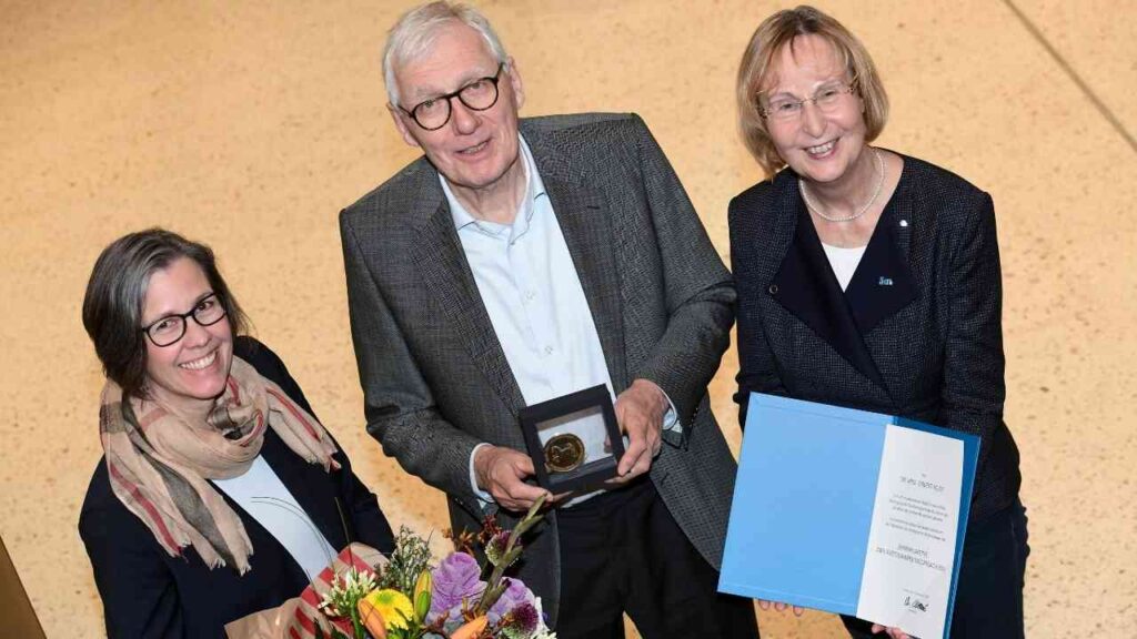 Drei Personen halten eine Ehrenplakette und Blumenstrauß in einer Innenumgebung.