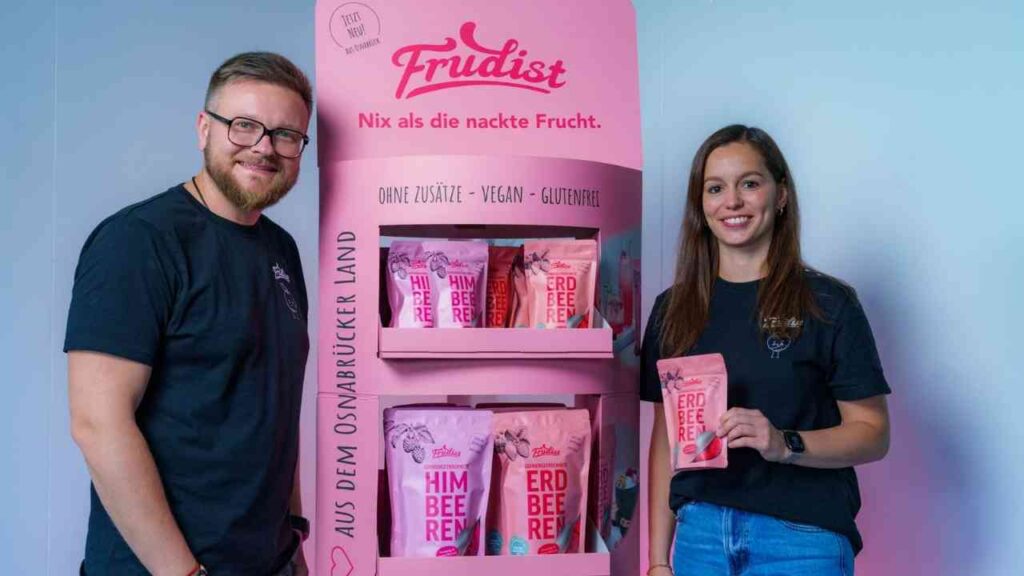 Zwei Personen stehen neben einem pinken Verkaufsdisplay mit Frudist-Produkten, die Himbeeren und Erdbeeren zeigen.