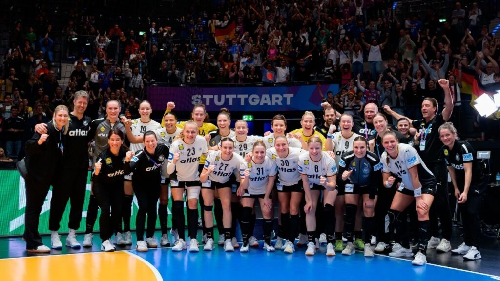 Handballmannschaft posiert nach dem Spiel in der Halle mit jubelnden Fans im Hintergrund.