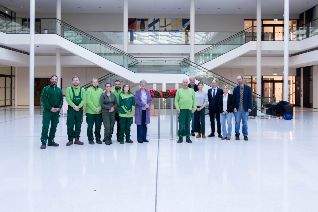 Gruppe von Personen in grüner Arbeitskleidung und Anzügen in einem modernen Innenraum mit Treppen und Pflanzen.
