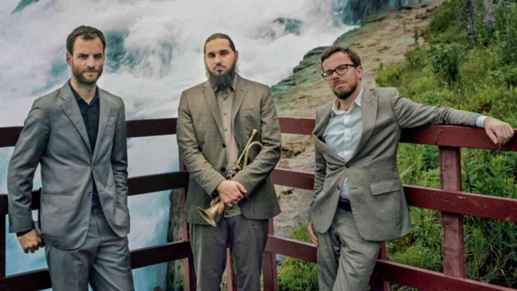 Jazztrio mit Trompete, bestehend aus drei Männern in Anzügen, posiert an einem Geländer mit Wasserfall im Hintergrund.