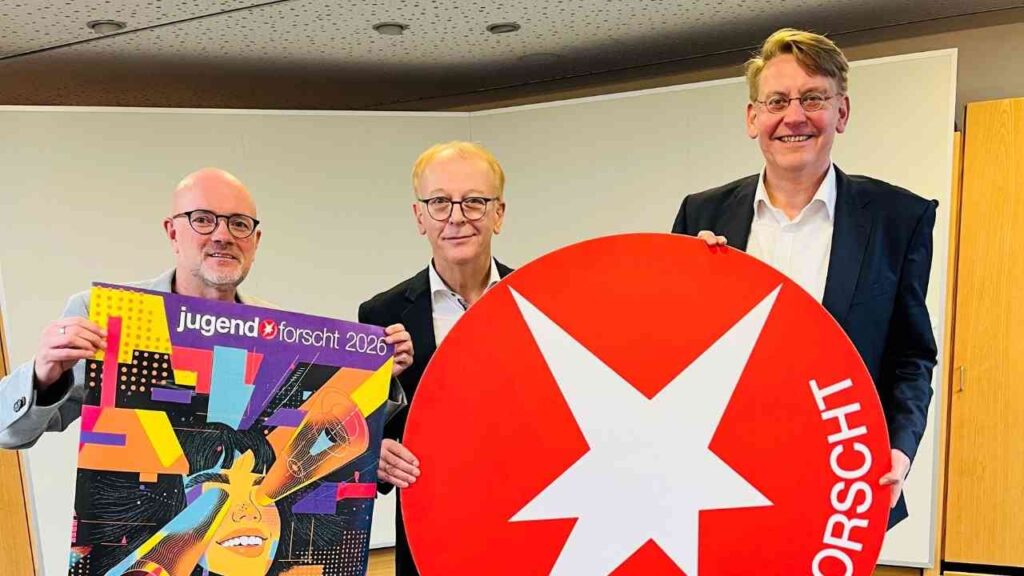 Drei Männer halten ein Plakat von 'Jugend forscht' und ein rotes Logo mit einem Stern in einem Besprechungsraum.