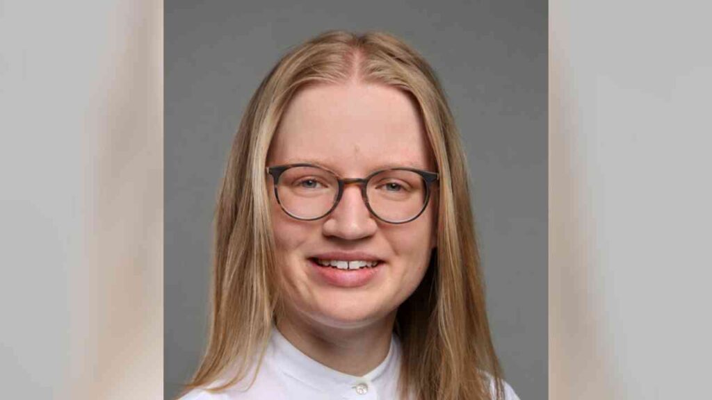 Porträt einer Frau mit langen, blonden Haaren und Brille, die in einem weißen Hemd lächelt.