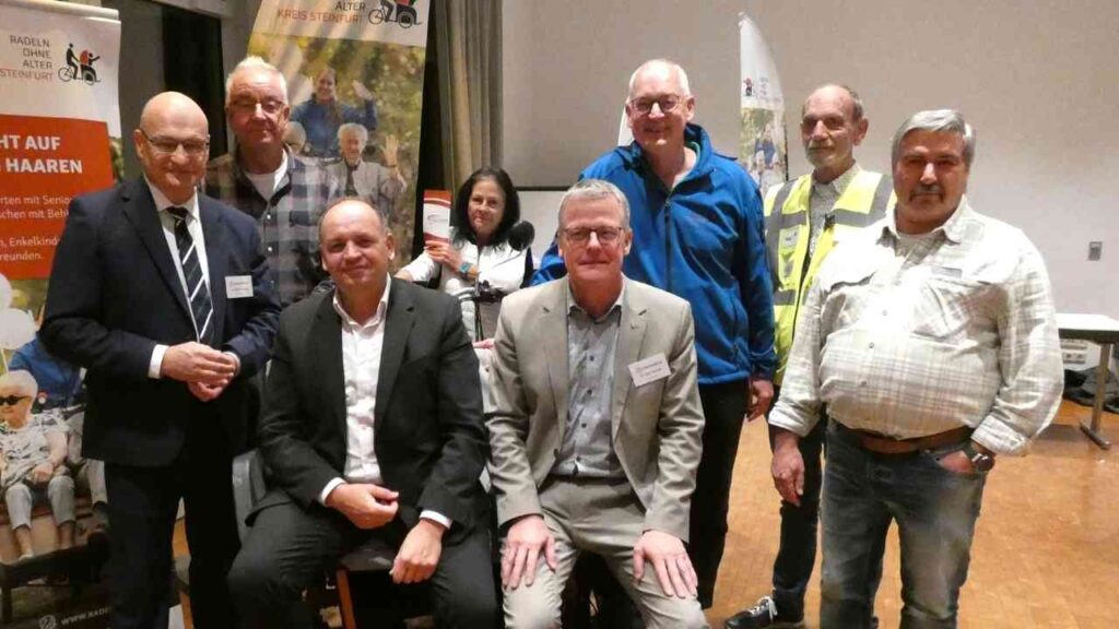 Gruppe von Personen, die an einer Veranstaltung teilnehmen, mit einem Banner im Hintergrund, das auf Rikscha-Fahrten hinweist.