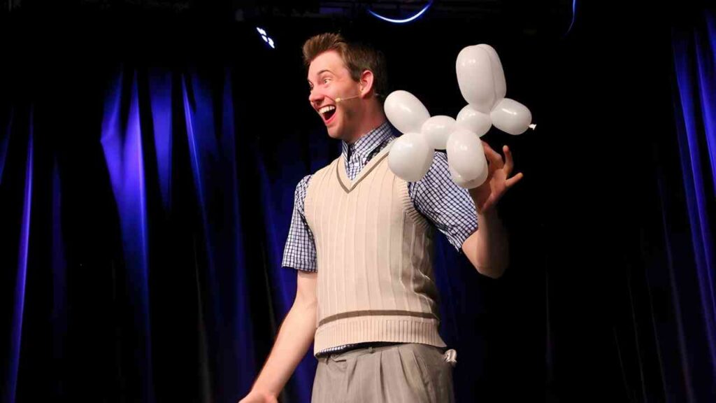 Ein Mann in einem gestreiften Pullover hält eine Ballonfigur in der Form eines Hundes während einer Aufführung.