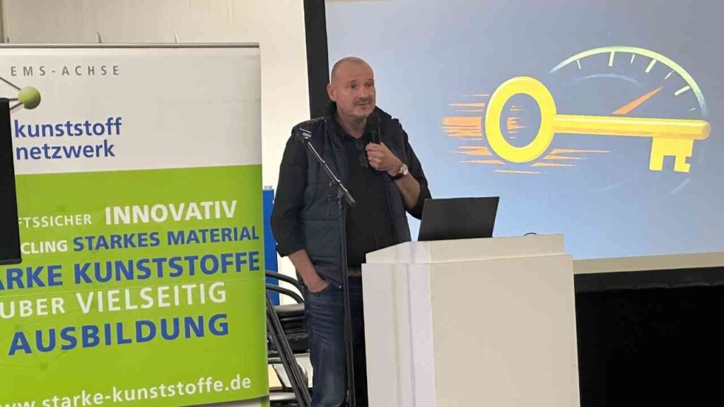 Mann spricht auf einer Veranstaltung mit einem Präsentationsbildschirm im Hintergrund, der einen Schlüssel und ein Geschwindigkeitsmesser zeigt.